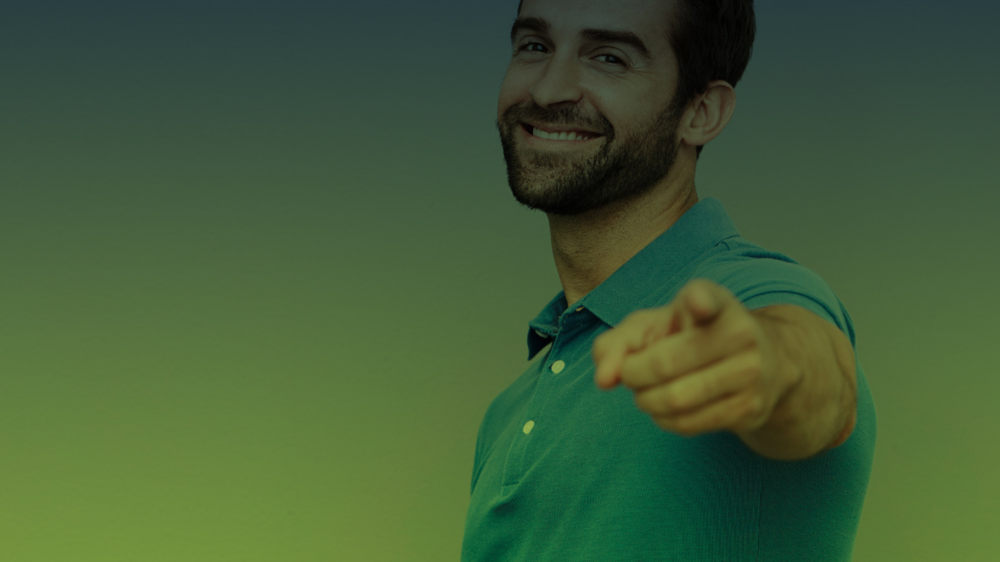 Homem com expressão confiante e sorriso no rosto aponta diretamente para a câmera, vestindo camisa polo verde.
