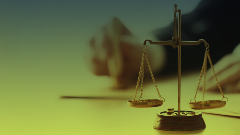 Close de uma balança dourada sobre uma mesa, símbolo da justiça, com mãos desfocadas ao fundo, simbolizando organização de classe.