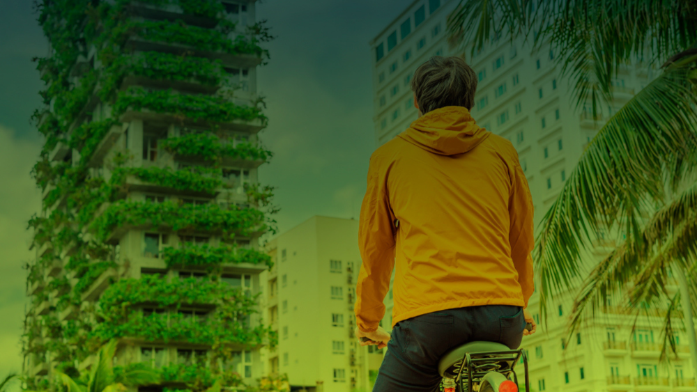 Homem de jaqueta amarela anda de bicicleta e observa prédio com fachada repleta de vegetação, destacando urbanismo sustentável.