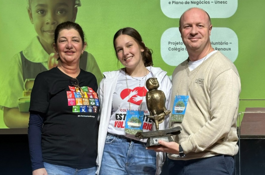 Na foto, representantes do projeto premiado na categoria Instituição de Ensino posam juntos no palco. Eles seguram o troféu, que é uma escultura de uma coruja. Todos sorriem para a câmera.