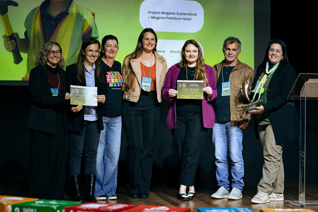 Na foto, representantes dos projetos finalistas e do projeto premiado do Fórum ODS Brasil 2025 posam juntos no palco. Duas pessoas seguram certificados de menção honrosa e uma pessoa segura o troféu, uma escultura de uma baleia-franca-austral. Todos sorriem para a câmera.