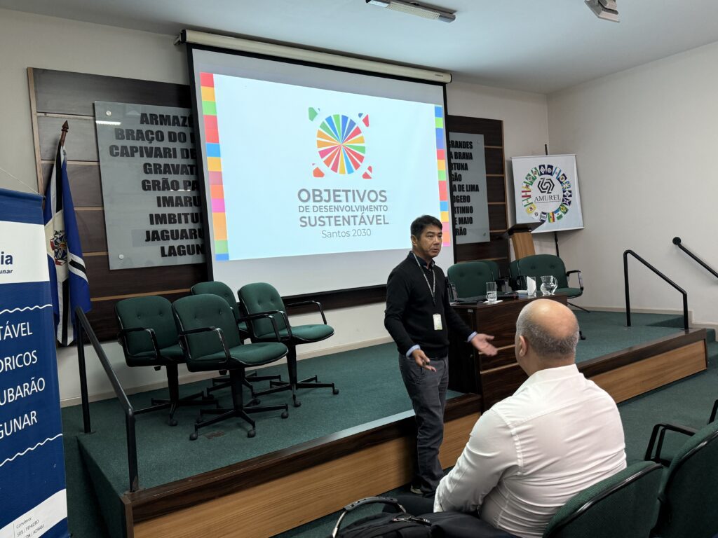 Homem em pé explica slide com gráfico comparando os Objetivos de Desenvolvimento do Milênio (ODM) e os Objetivos de Desenvolvimento Sustentável (ODS), diante de público atento.