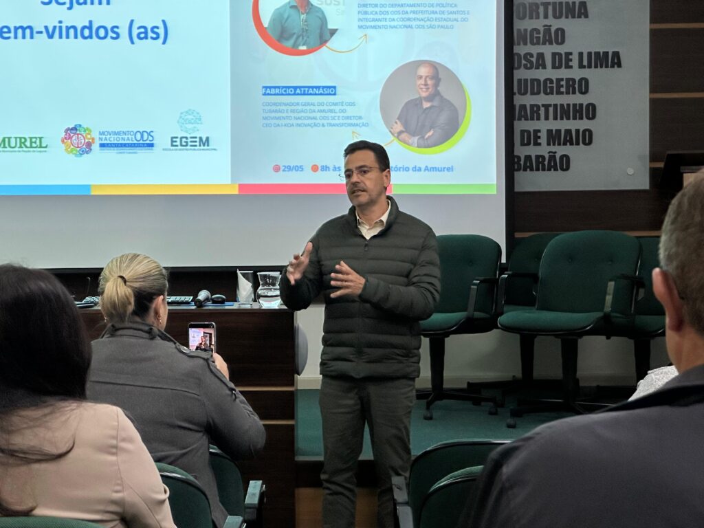 Homem em pé explica slide com gráfico comparando os Objetivos de Desenvolvimento do Milênio (ODM) e os Objetivos de Desenvolvimento Sustentável (ODS), diante de público atento.