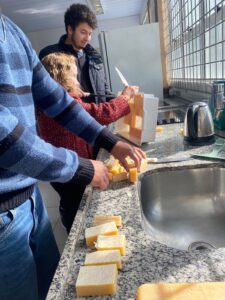 Três pessoas trabalham em uma bancada de cozinha. Uma mulher, de suéter vermelho, corta uma grande peça amarela em barras menores, enquanto um homem jovem observa. Na bancada de granito, várias barras retangulares de sabão amarelo já estão cortadas e alinhadas ao lado da pia de inox. Ao fundo, há uma janela gradeada deixando entrar luz natural e eletrodomésticos como chaleira elétrica.