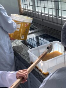 Pessoa com jaleco branco segura um balde grande amarelo da marca Soya, despejando uma mistura pastosa de cor amarelada em um recipiente plástico retangular sobre uma bancada de granito. Outra pessoa segura uma haste de madeira usada para mexer a mistura. Ao fundo, há um fogão elétrico e uma janela gradeada que deixa entrar luz natural.