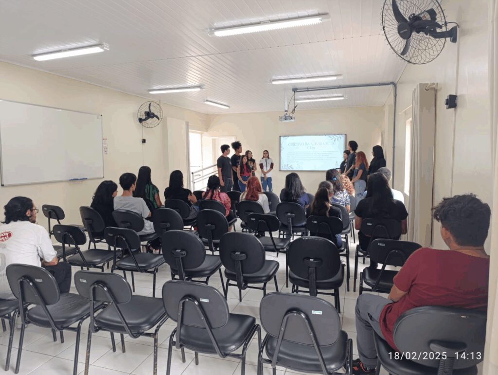 Sala de aula clara, com paredes brancas, ventiladores de parede e cadeiras pretas dispostas em fileiras. Cerca de quinze alunos estão sentados, de costas para a câmera, assistindo a uma apresentação feita por oito jovens que estão em pé, próximos a um projetor. Na tela projetada lê-se “OBJETIVO DA ATIVIDADE NO CEJA”. A foto tem marcação de data e hora no canto inferior direito: 18/02/2025 às 14:13.