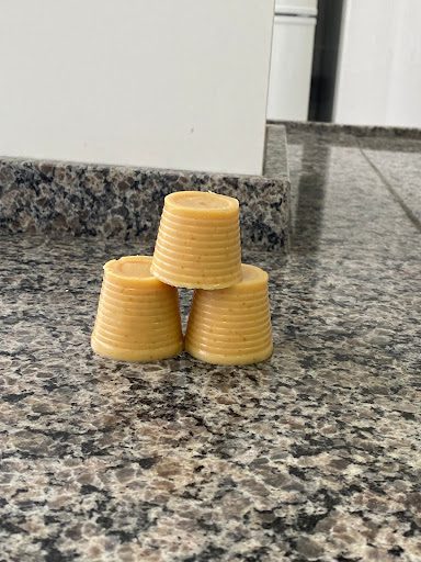 Três pedaços de sabão artesanal, em formato cilíndrico e cor amarela, empilhados em forma de pirâmide sobre uma bancada de granito cinza. Ao fundo, parte de uma parede e móveis brancos.