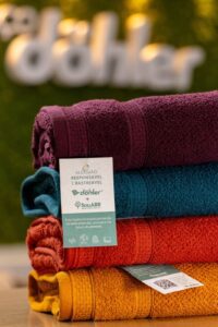 Pilha de quatro toalhas coloridas (bordô, azul, laranja e mostarda) com etiqueta destacando: “Algodão Responsável e Rastreável – Döhler + SouABR”. O texto diz que a toalha foi criada pensando no bem-estar das pessoas e no futuro do planeta. Ao fundo, o logo da Döhler aparece desfocado sobre um painel verde.