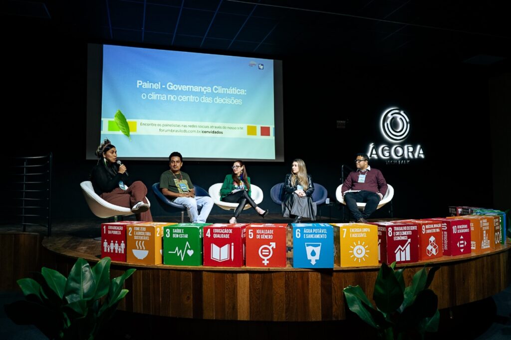 Painel "Governança Climática: o clima no centro das decisões" no Fórum Brasil ODS 2025. Cinco painelistas sentados no palco do Ágora Tech Park debatem o tema. À frente, caixas representando os ODS da ONU, como Erradicação da Pobreza, Energia Limpa e Igualdade de Gênero.