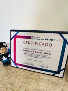 Certificado do Prêmio Ailema Pucú emoldurado, concedido a Jamile de Souza Lima pelo Conselho Federal de Administração, apoiado sobre uma superfície clara. Ao lado do certificado há um pequeno boneco decorativo.