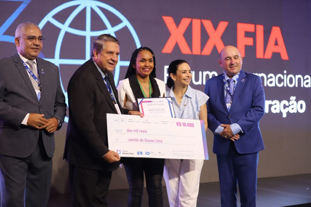 Jamile Lima, mulher negra, sorri enquanto segura um certificado e um cheque simbólico durante cerimônia de premiação no XIX Fórum Internacional de Administração. Ela está ao lado de representantes da organização, em um palco com painel ao fundo identificando o evento.