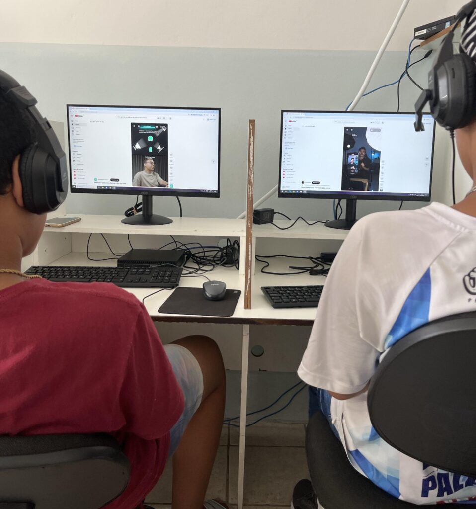 Duas pessoas, vistas de costas e usando fones de ouvido, estão sentadas diante de computadores. Nas duas telas aparecem vídeos abertos em uma plataforma online.