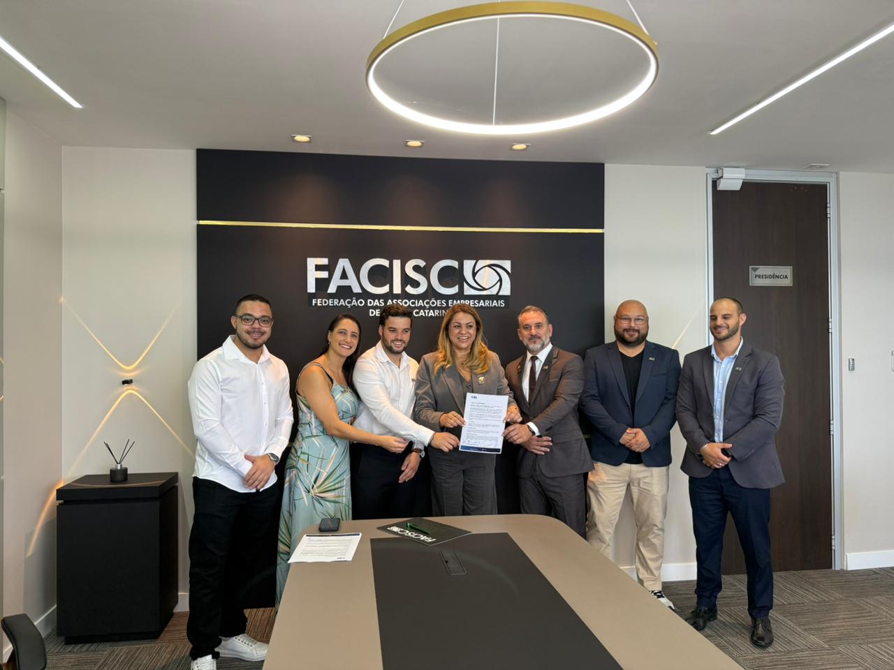 Grupo de sete pessoas posa com documento durante encontro na sede da FACISC relacionado ao DEL Habitação em Içara.